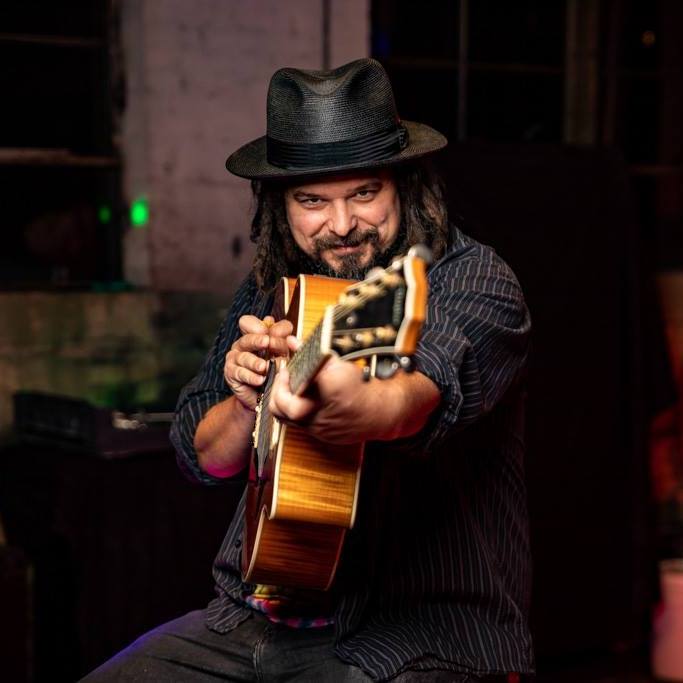 Musician, Michael Doussan, wearing top hat, holding a guitar pointed at the camera